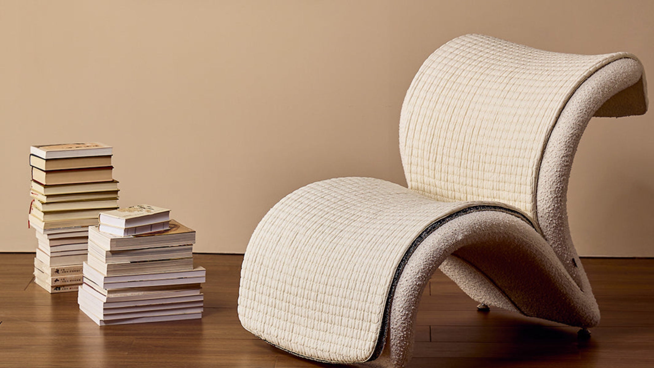 Modern beige chair against a beige wall with a stack of books and a brown roll of paper.